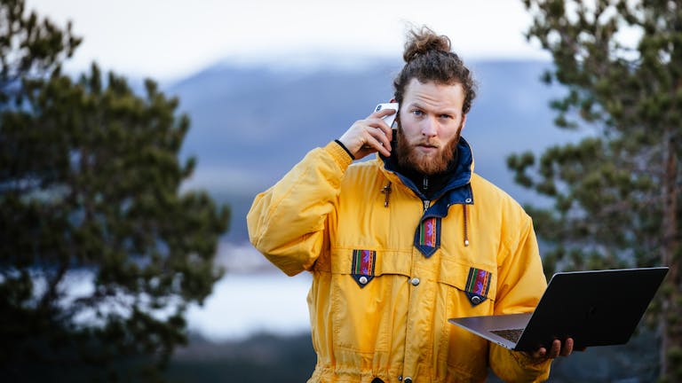 Man managing work remotely with laptop and smartphone amidst Norway's natural beauty.